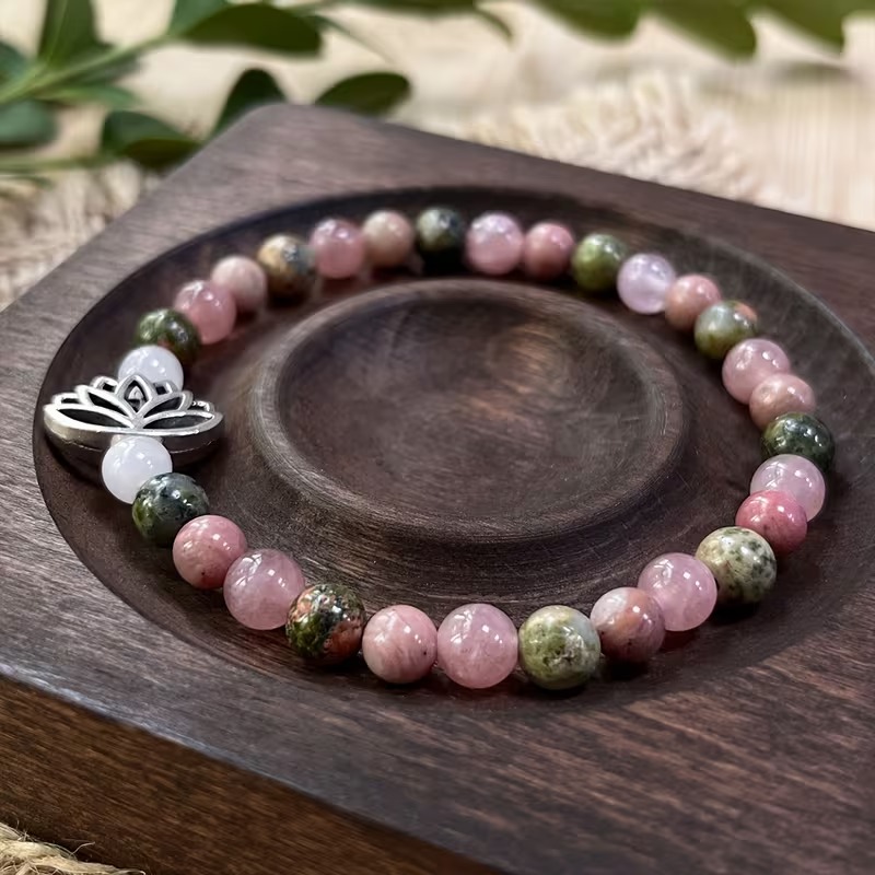 Armbånd med rosa og grønne perler og sølvfarvet lotusmotiv på en mørk træbakke.